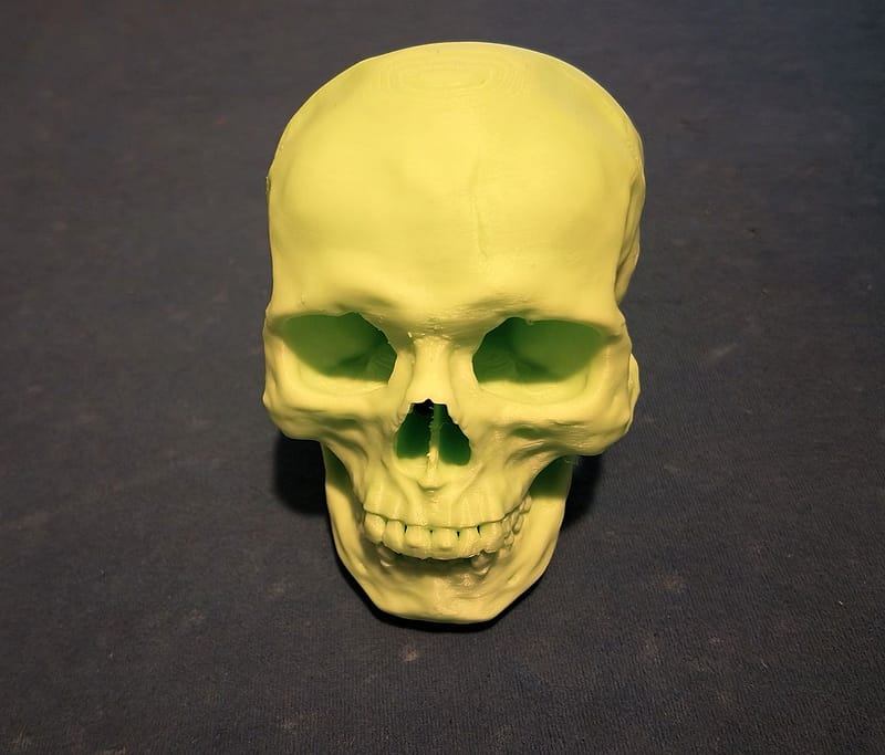 Lichtgroene, 3D-geprinte schedel van voren op een blauwe ondergrond, met diepe oogkassen, een neusopening en een open kaak met tanden