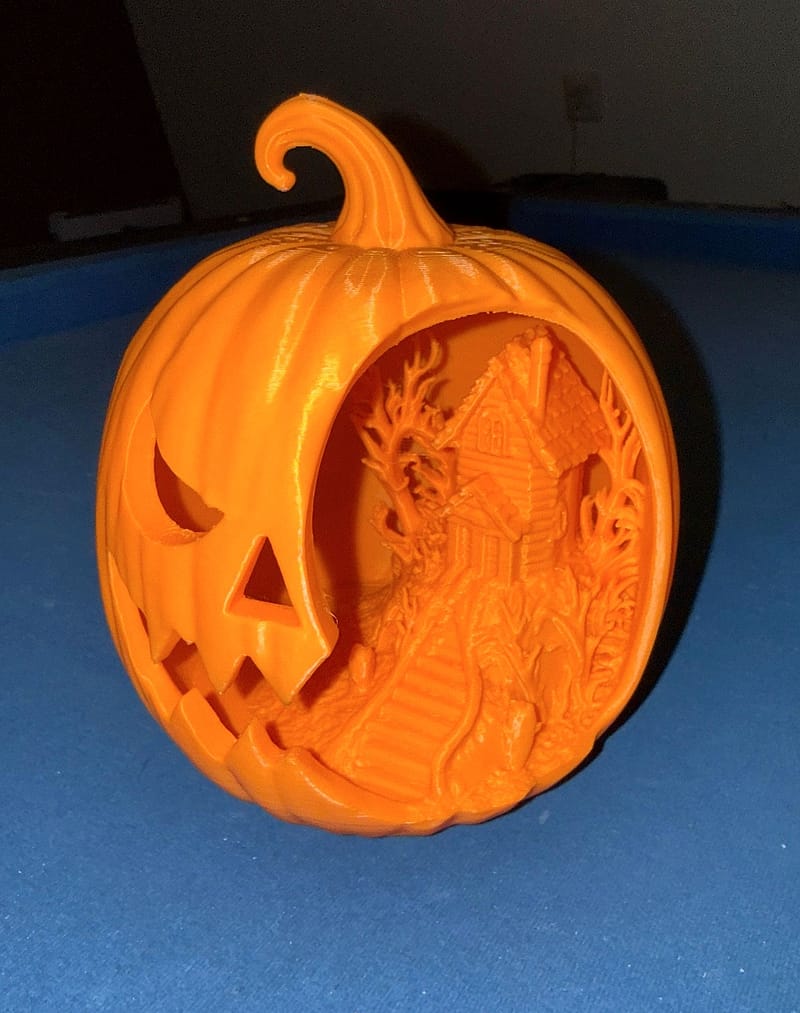 Oranje, 3D-geprinte Halloween pompoen schuin van voren: links het uitgesneden gezicht, rechts de open zijde met een spookhuis-diorama met een huis, kale bomen en een trap