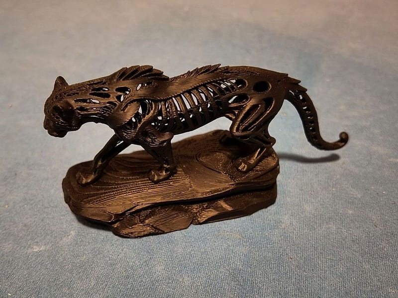 Zwarte, 3D-geprinte pantersculptuur van opzij op een rotsachtige basis; het lichaam is opengewerkt met zichtbare ribben en de staart krult omhoog